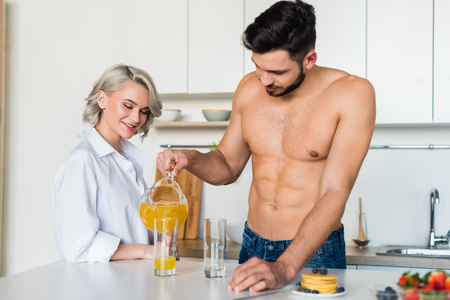 shirtless young man pouring juice to smiling girlfriend standing near by in kitchenの写真素材