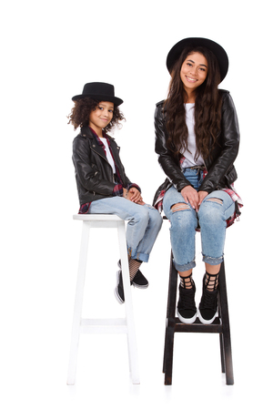 Stylish mother and daughter in hats and leather jackets sitting on chairs isolated on whiteの写真素材