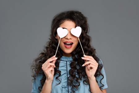 Excited young woman covering eyes with hearts on sticks isolated on greyの写真素材