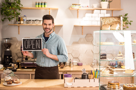 Smiling barista with open blackboard in hands at counter in coffee shop