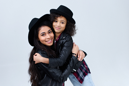 Smiling stylish mother and daughter in similar clothes embracing and looking at camera isolated on greyの写真素材