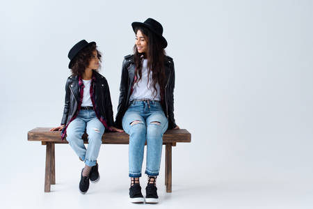 Happy mother and daughter in similar clothes sitting on bench together isolated on greyの写真素材