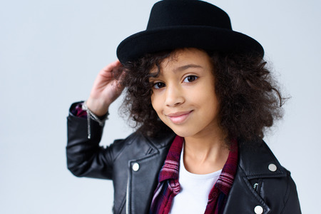 Close-up portrait of adorable little child in leather jacket and hat looking at camera isolated on greyの写真素材
