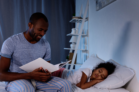 african american father reading book for sleeping daughter at homeの写真素材