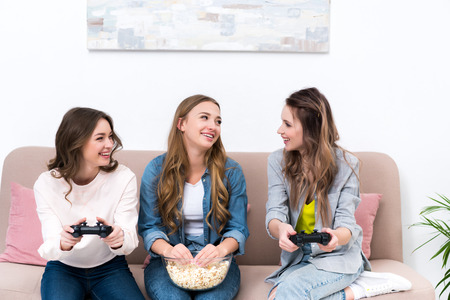 beautiful young girlfriends eating popcorn and playing with joysticks together at homeの写真素材