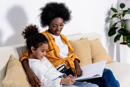 African american mother and daughter reading book on sofa at homeの写真素材