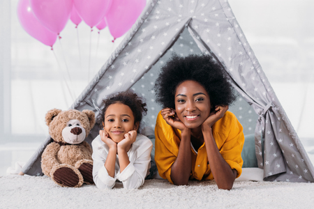 african american mother and daughter resting chins on hands and looking at camera at homeの写真素材