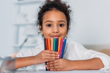 Adorable African american kid holding colored felt pens and looking at camera at homeの写真素材