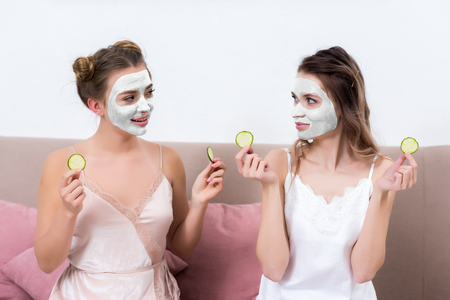Beautiful girls in pajamas and facial masks holding slices of cucumber and looking at each otherの写真素材