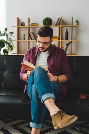 Young male in glasses sitting on leather couch and reading bookの写真素材