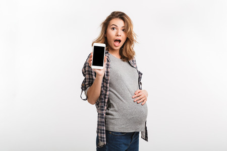 portrait of shocked pregnant woman showing smartphone with blank screen isolated on greyの写真素材