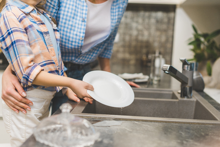 partial view of little daughter helping mother to wash dishes after dinner at home