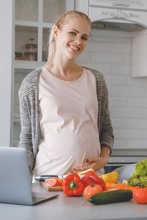 smiling pregnant woman standing near vegetables in kitchenの写真素材