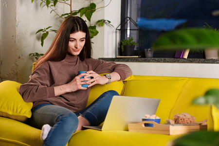 portrait of woman with cup of coffee using laptop on sofa at homeの写真素材