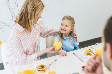 mother giving apple to daughter during breakfast at homeの写真素材