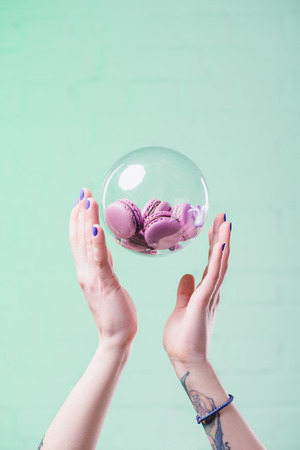 cropped shot of woman throwing up glass ball with delicious macarons on turquoiseの写真素材