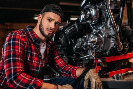 handsome bike repair station worker with wrench sitting in front of motorcycle and looking at cameraの写真素材