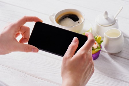 cropped shot of woman with smartphone with blank screen, cup of coffee, jag of cream and sweet muffin on wooden surfaceの写真素材