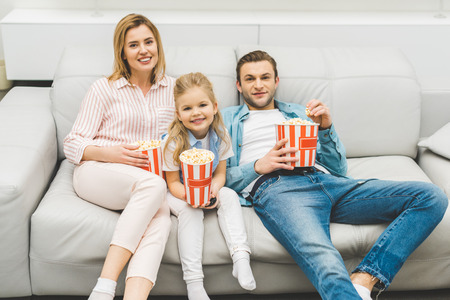 smiling family with popcorn watching film together at homeの写真素材