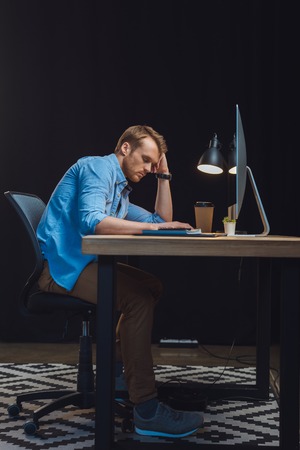 young overworked businessman sleeping at table with desk lamp, coffee cup and computer in officeの写真素材