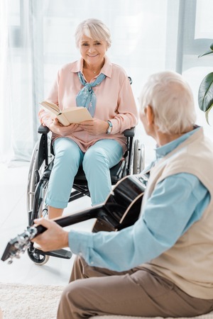 senior woman sitting in wheelchair and reading book while senior man playing acoustic guitarの写真素材