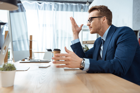 side view of handsome businessman in eyeglasses gesturing by hands at table with computer monitor in officeの写真素材