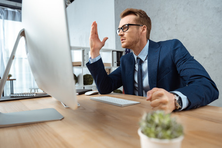 irritated businessman in eyeglasses gesturing by hand at table with computer monitor in office