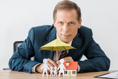 businessman with paper cut family, house model and umbrella on wooden tableの写真素材