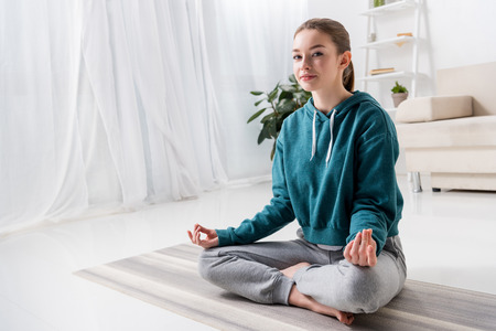 beautiful girl sitting in lotus position on yoga mat and looking at camera at home