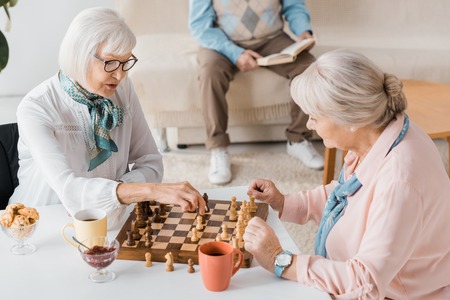 senior women playing chess and drinking coffee while man reading book on sofaの写真素材