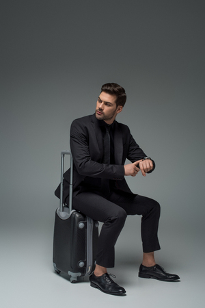 Business tourist sitting on suitcase and pointing at wristwatch on greyの写真素材