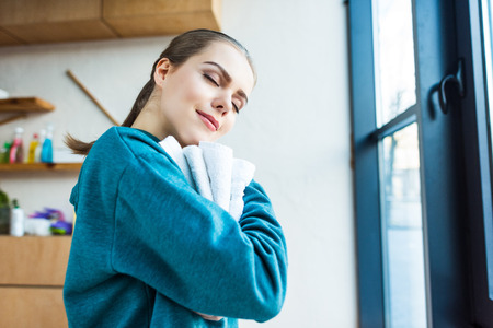 smiling young woman with closed eyes hugging clean towels at homeの写真素材