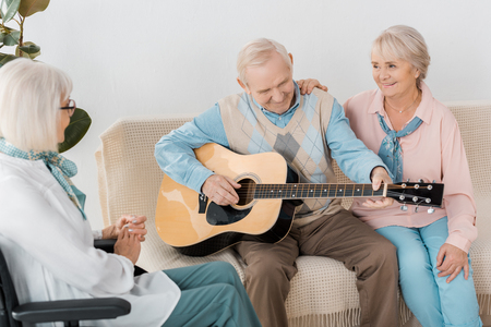 senior man sitting on sofa and playing acoustic guitar for womenの写真素材