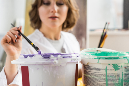 Young creative girl dunks a paintbrush into the paint in light studioの写真素材