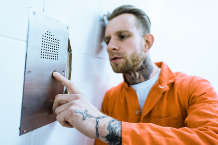 Tattooed criminal in orange uniform pressing button in prison cellの写真素材