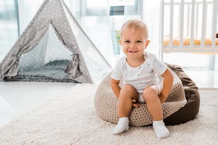 Adorable smiling toddler sitting on bean bag chair and looking at camera in nursery roomの写真素材