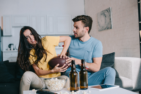 Angry young woman fighting for american football ball with husband during match on tv at homeの写真素材