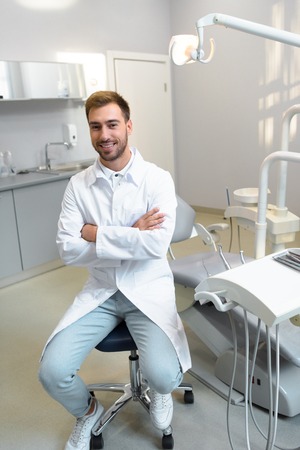 Handsome young dentist with crossed arms looking at camera while sitting at officeの写真素材
