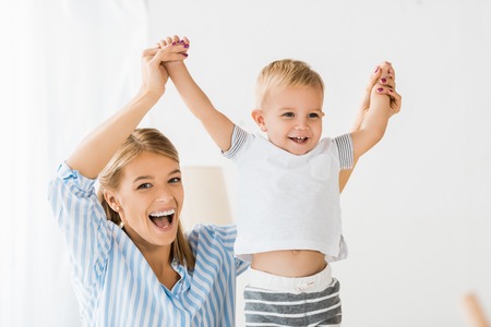 Cheerful mother looking at camera and holding smiling toddler in hands
