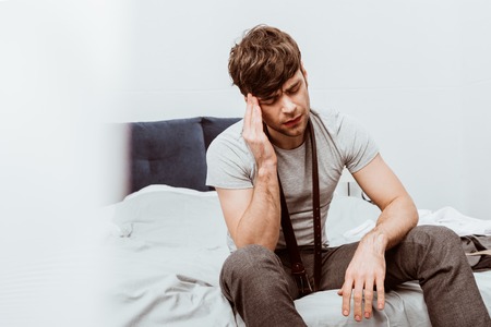 Handsome young businessman with belt over neck sitting on bed and having headache at home