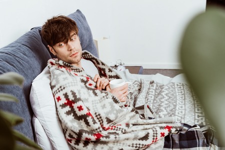 Selective focus of sick man wrapped in blanket sitting with cup of tea in bed at homeの写真素材