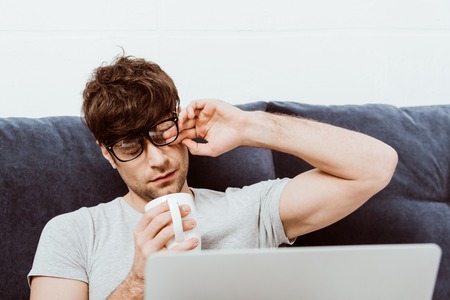 Tired male freelancer in eyeglasses holding coffee cup and sitting on bed with laptop at homeの写真素材