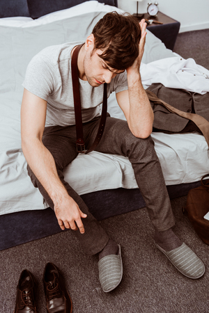 Young businessman with belt over neck sitting on bed and having headache at homeの写真素材