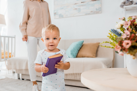 Adorable toddler holding book and looking at camera indoors with mother on backgroundの写真素材