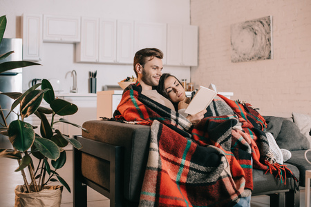 Smiling young couple reading book together on couch under plaid at homeの写真素材
