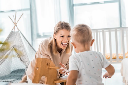 Happy mother and toddler playing with toy wooden rocking horse chair in nursery roomの写真素材