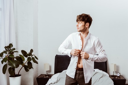 Side view of handsome businessman buttoning up white shirt in bedroom at homeの写真素材