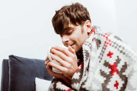 Sick man wrapped in blanket drinking tea in bed at homeの写真素材