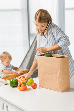 mom holding groceries while talking on smartphone with toddler in baby chair on backgroundの写真素材
