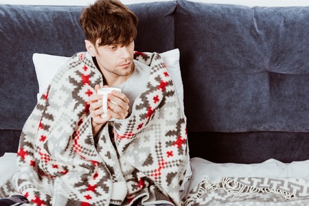 high angle view of sick man wrapped in blanket sitting with cup of tea in bed at homeの写真素材